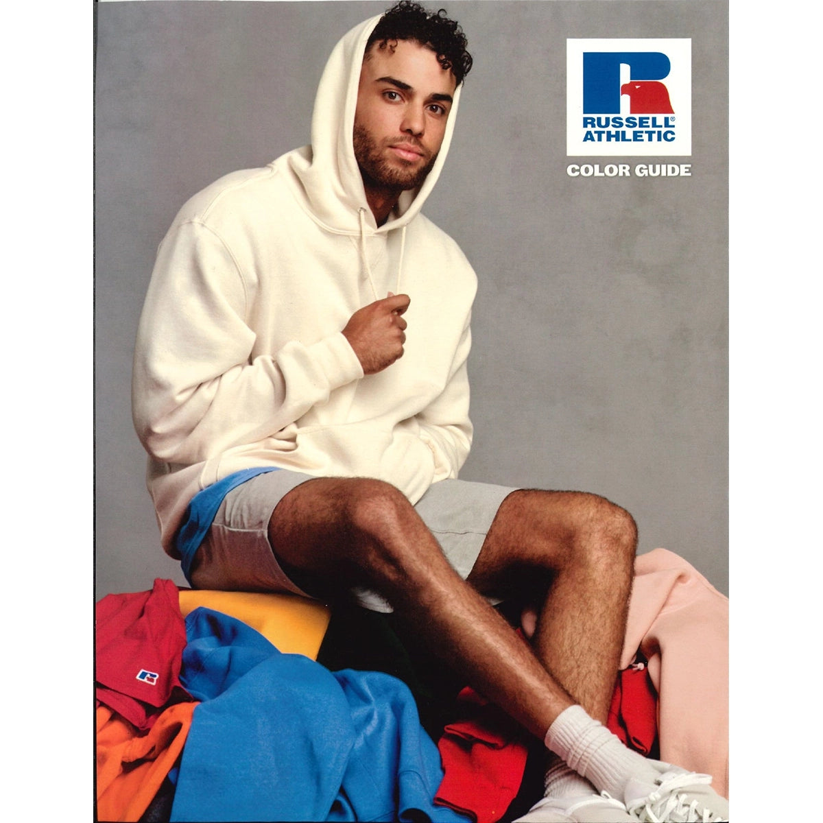 A man wearing a white Russell Athletic hoodie sitting on a pile of colorful apparel for the 2025 Swatch Card.