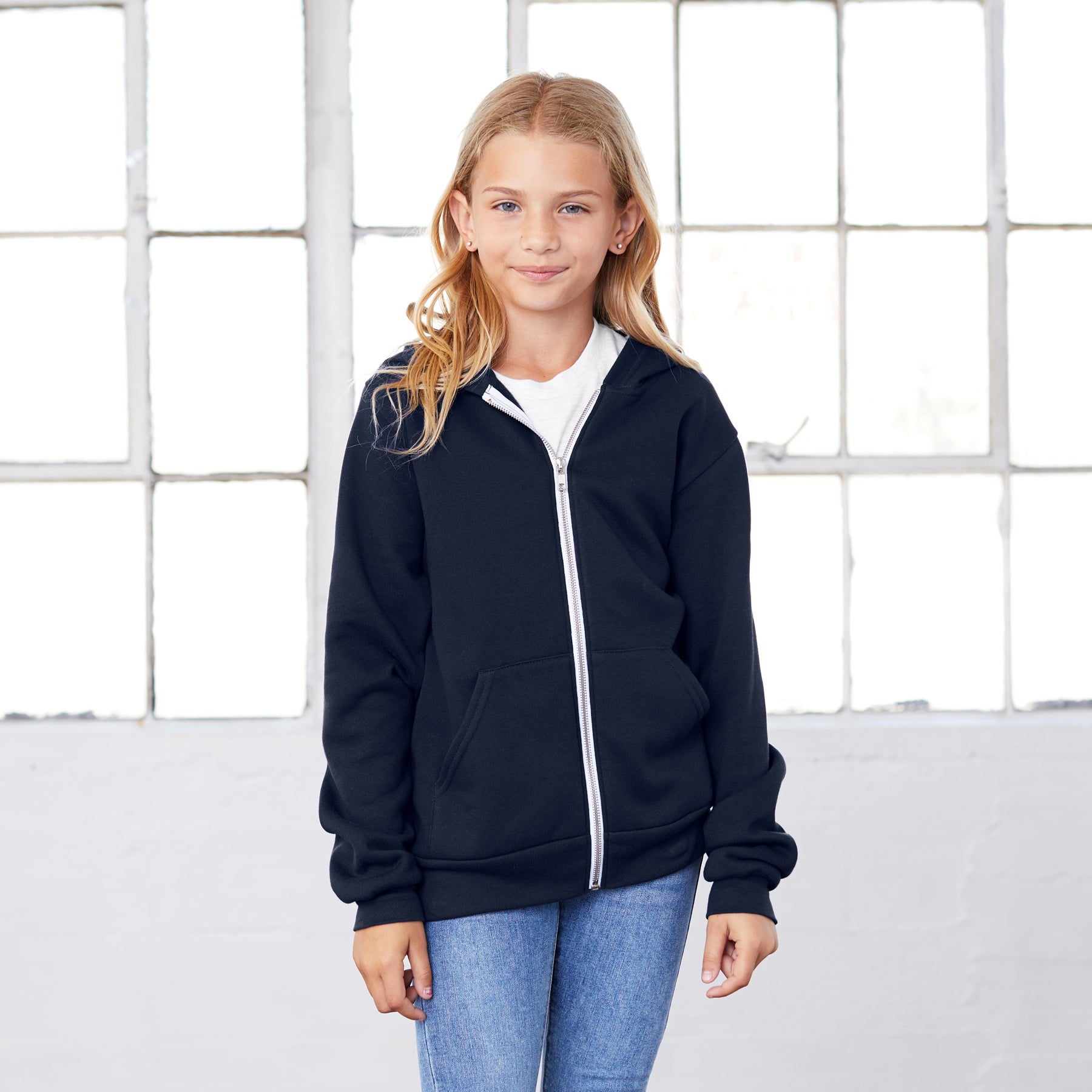 A young girl wearing a navy Bella+Canvas Youth Poly-Cotton Fleece Full-Zip Hoodie over a white shirt.
