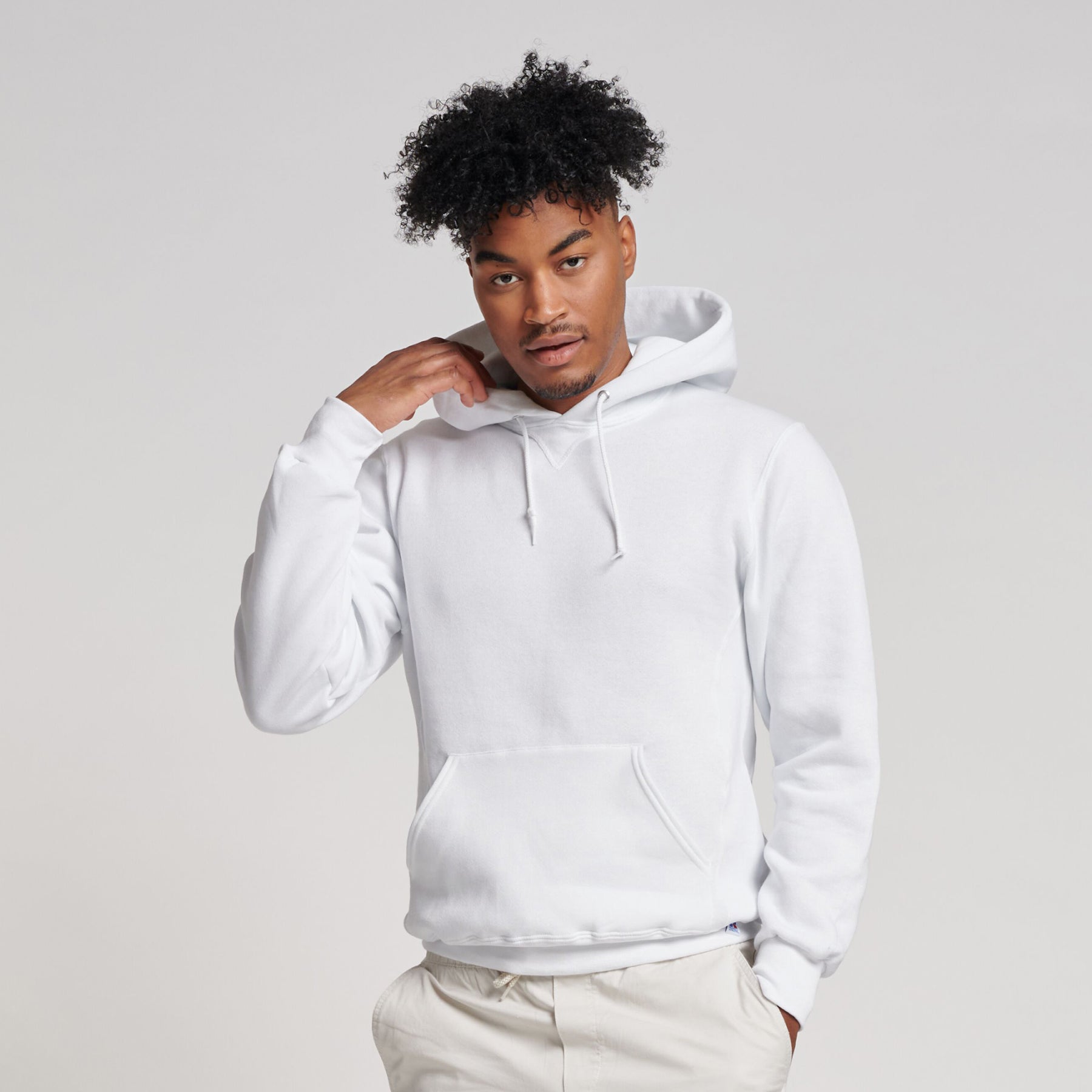 A man with curly hair wearing a white Russell Athletic DRI-POWER Adult Fleece Unisex Hoodie against a gray background.