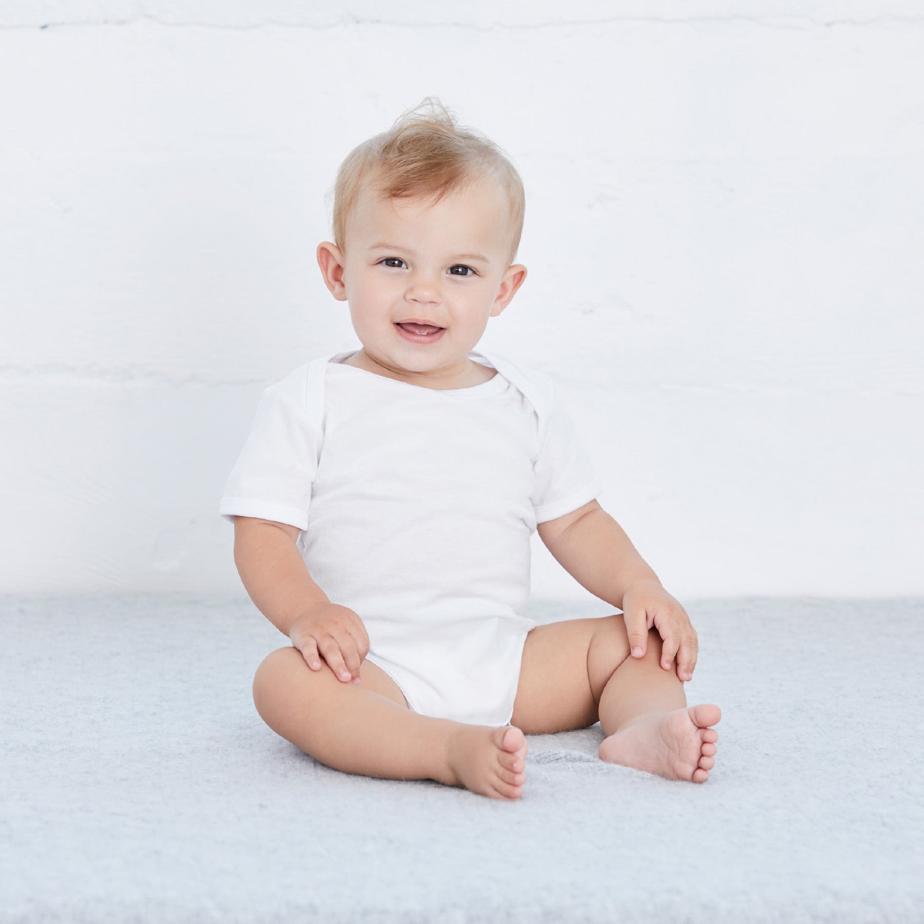 A happy infant sitting on a gray surface wearing a white Bella+Canvas Infant Jersey Short Sleeve One Piece.