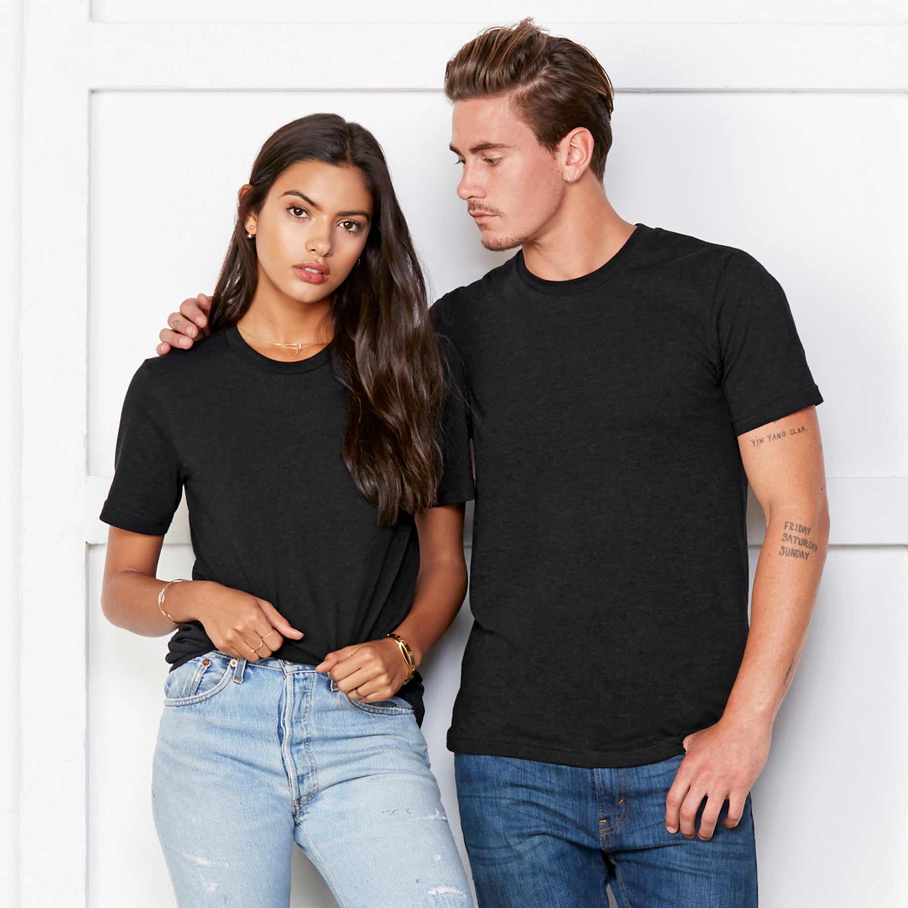 A man and woman wearing black heather Bella+Canvas Unisex Heather CVC Short Sleeve T-shirts posing against a white wall.