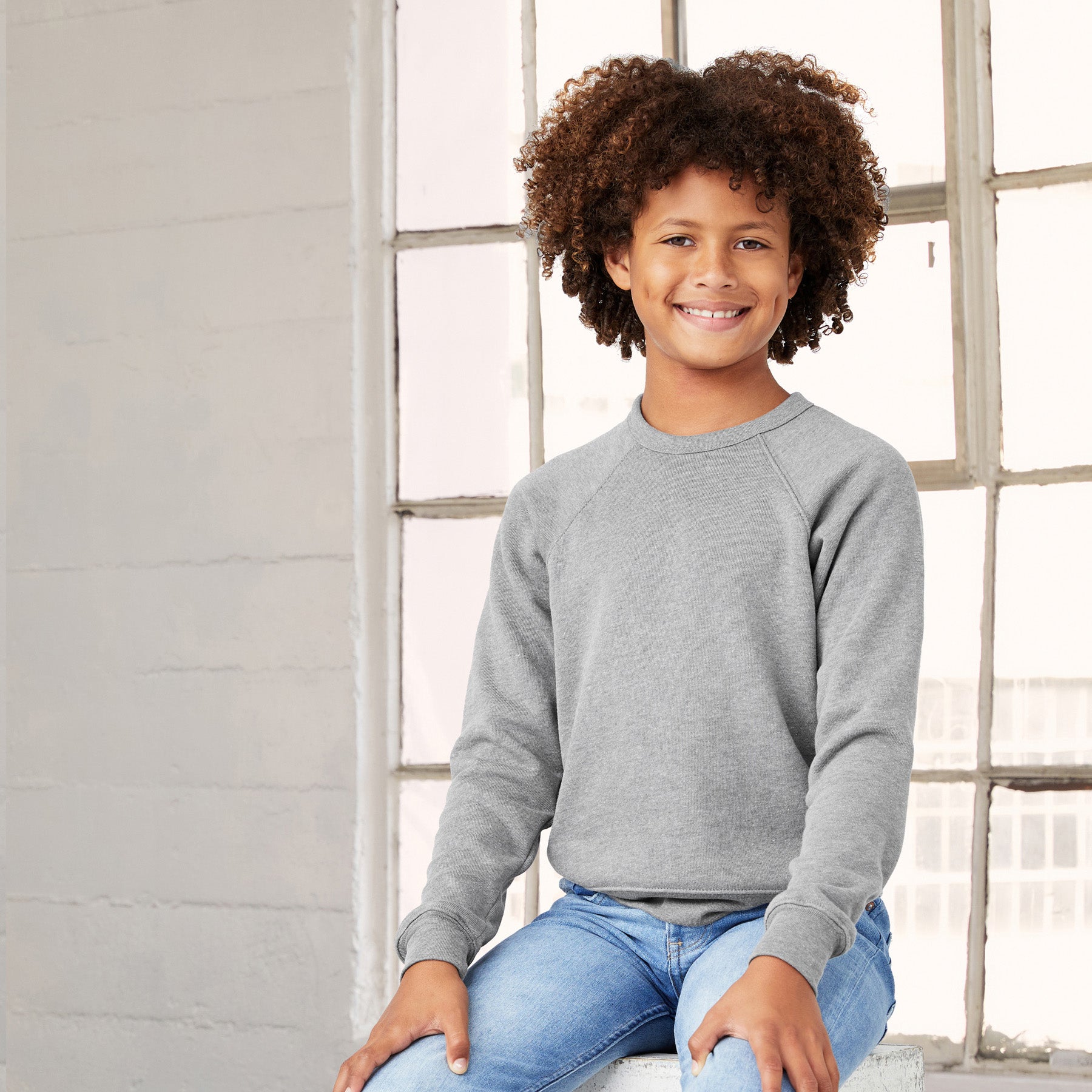 A child with curly hair wears a Bella+Canvas Youth Poly-Cotton Fleece Raglan Sweatshirt in Deep Heather.