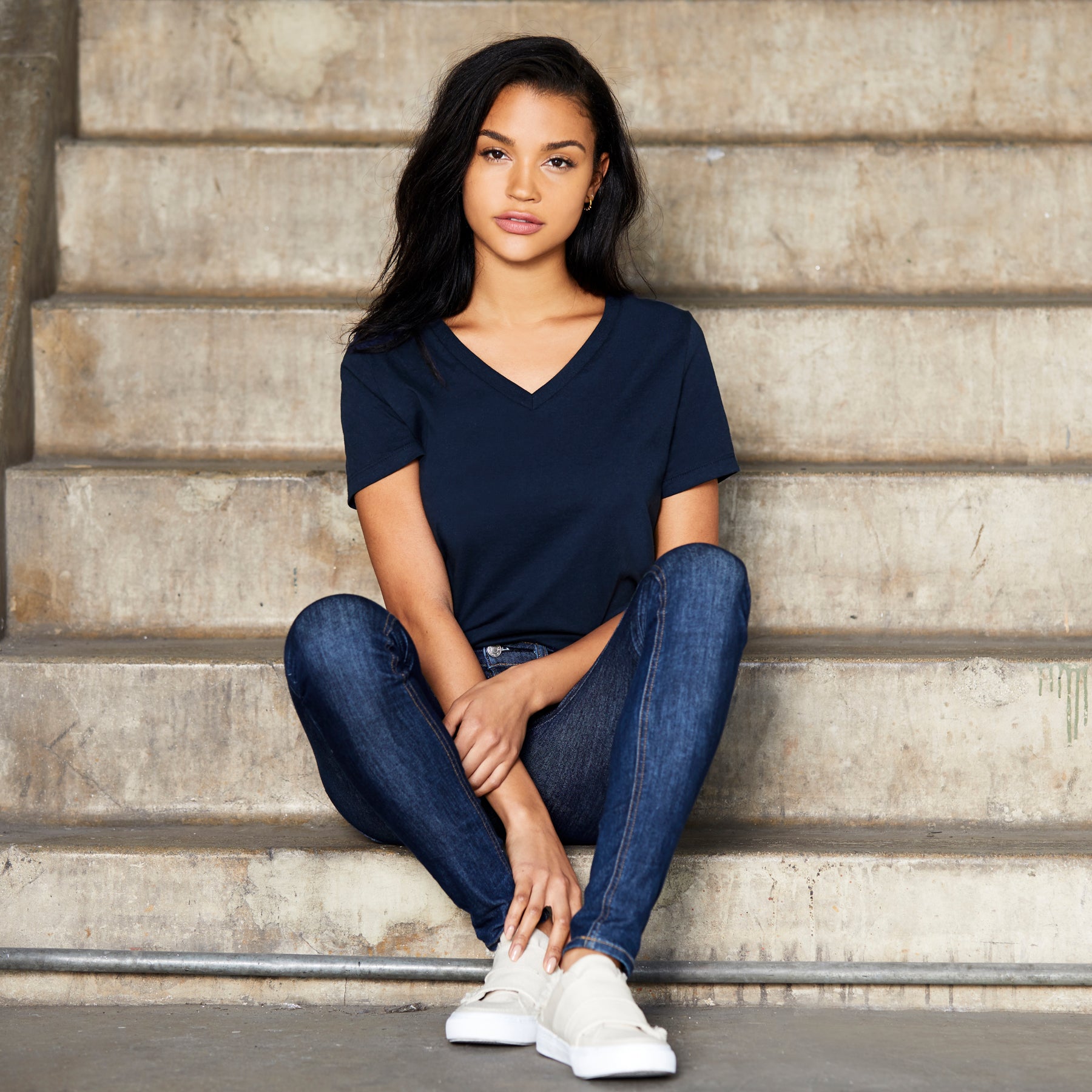 A woman wearing a navy Bella+Canvas Women's Relaxed Jersey Short Sleeve V-Neck Tee while sitting on concrete stairs.