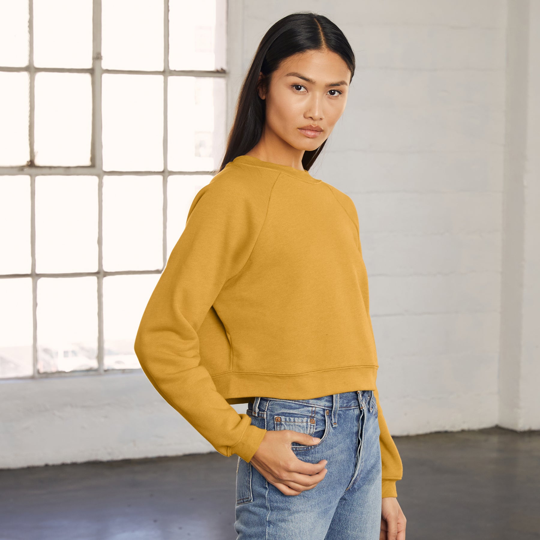 A woman wearing a mustard heather Bella+Canvas Women's Poly-Cotton Fleece Raglan Pullover with blue jeans.