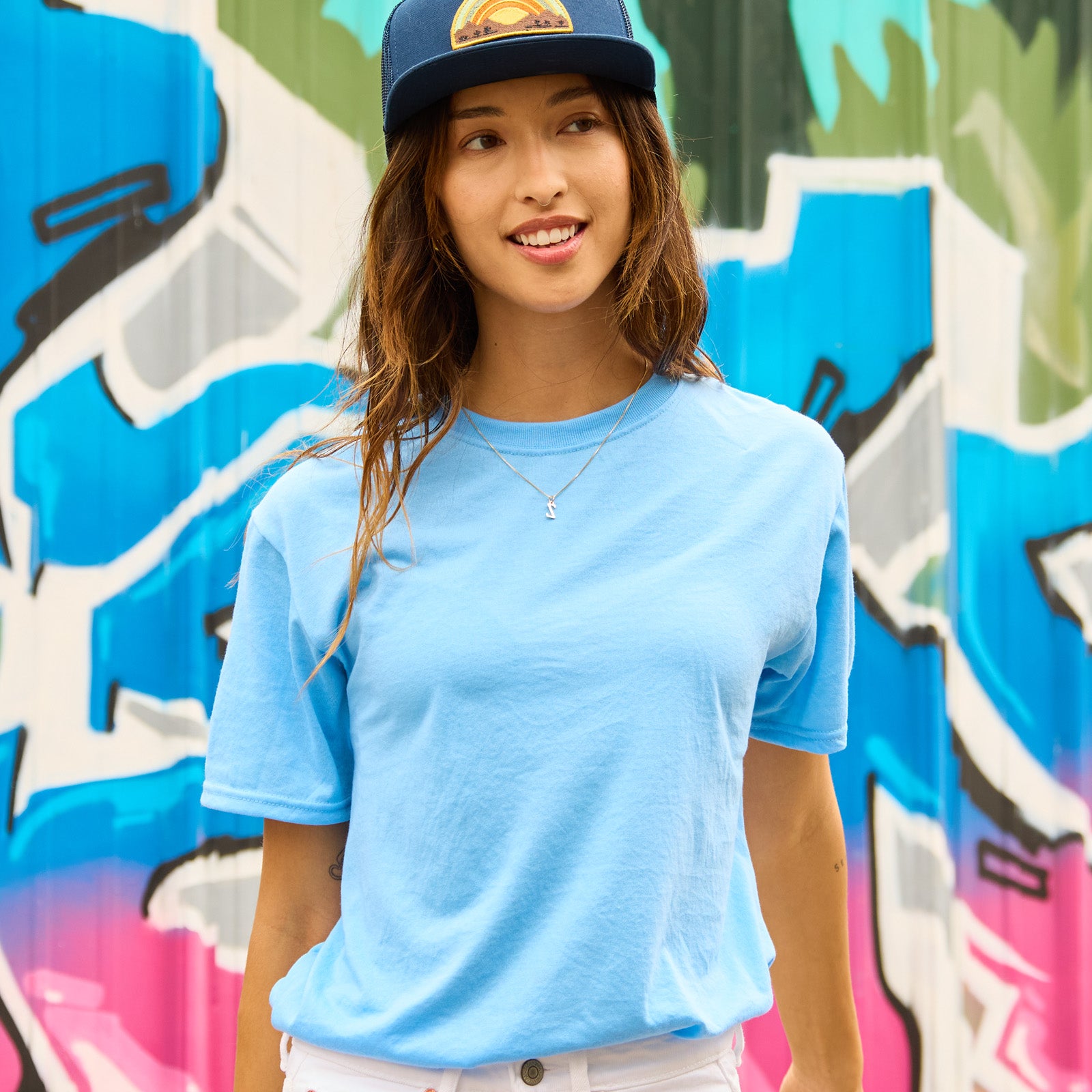 A woman wearing a light blue Jerzees Dri-Power unisex t-shirt and a baseball cap against a colorful graffiti background.