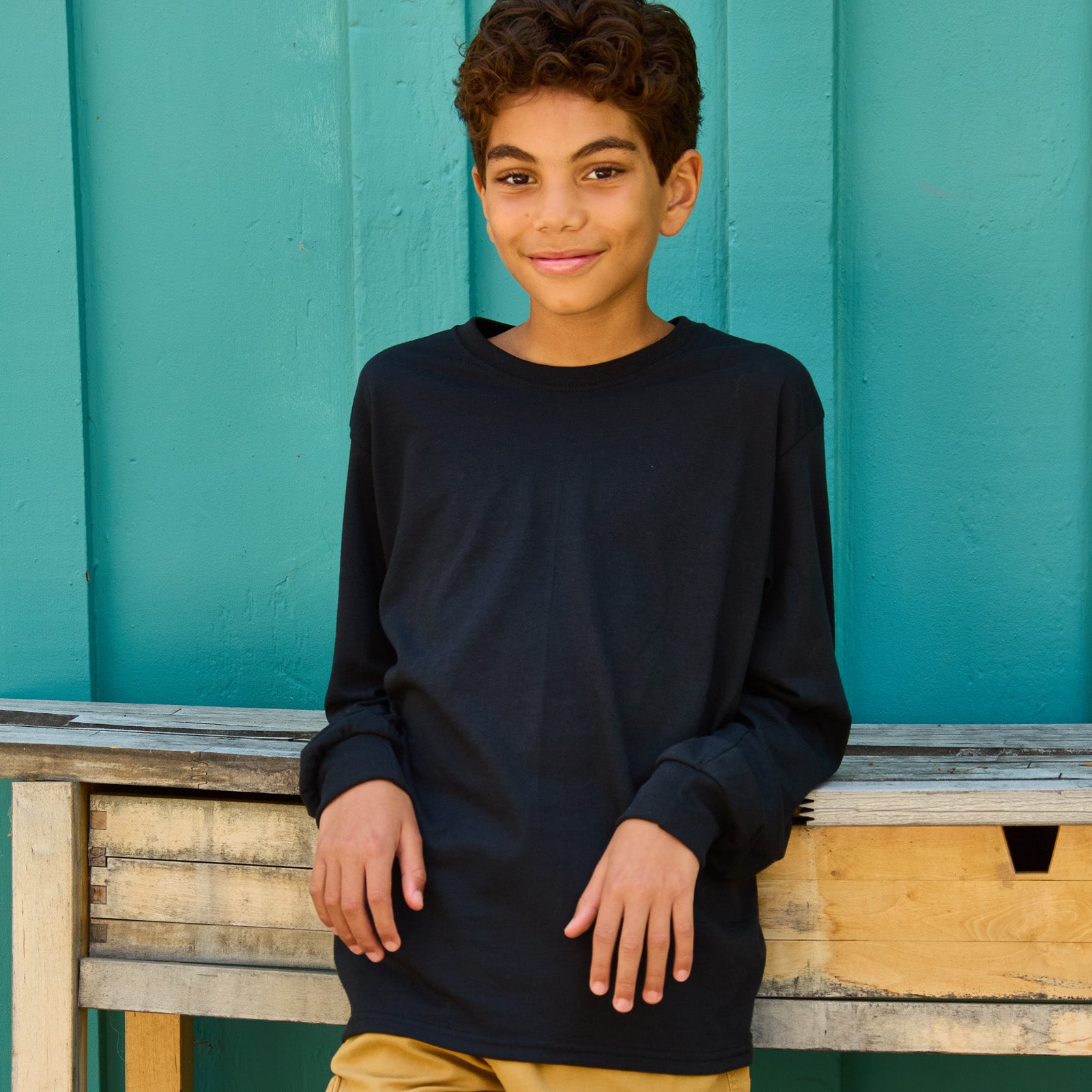 A young boy wearing a black Jerzees Dri-Power youth long-sleeve T-shirt while leaning against a wooden table.