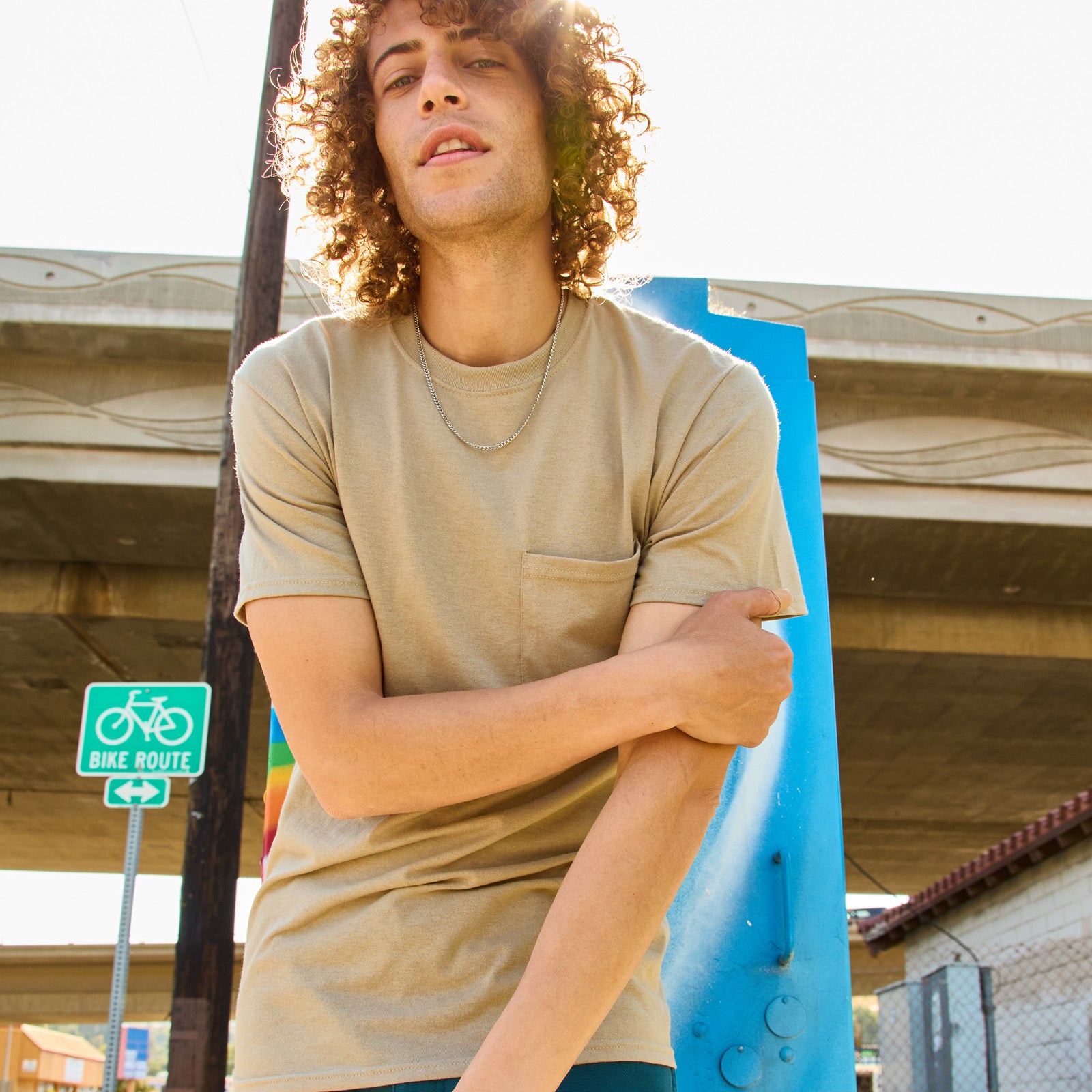 Model wearing a Jerzees Dri-Power Unisex Pocket T-Shirt in khaki, standing outdoors under a bridge.