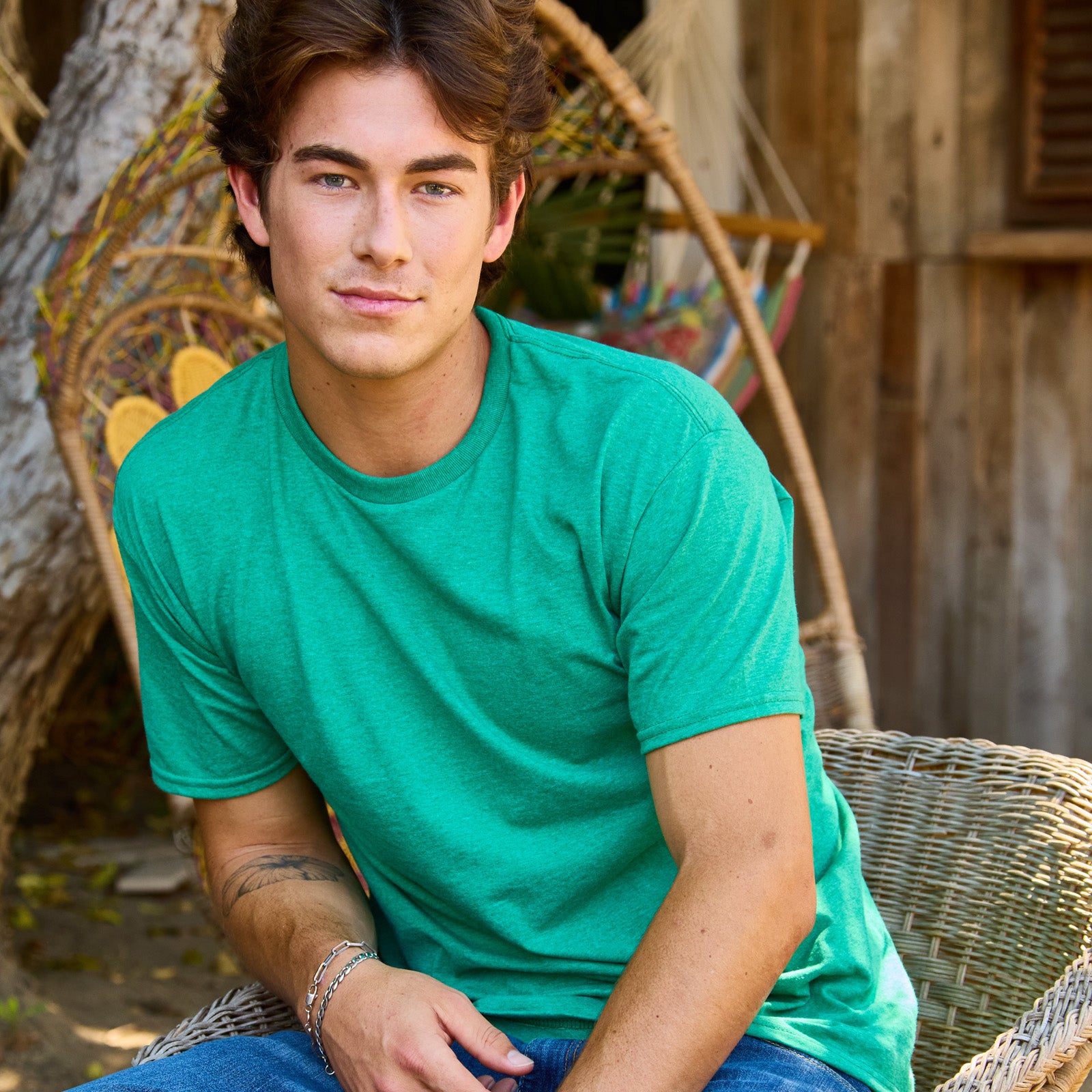A young man wearing a Jerzees Classics unisex t-shirt in retro heather green while sitting in a wicker chair.