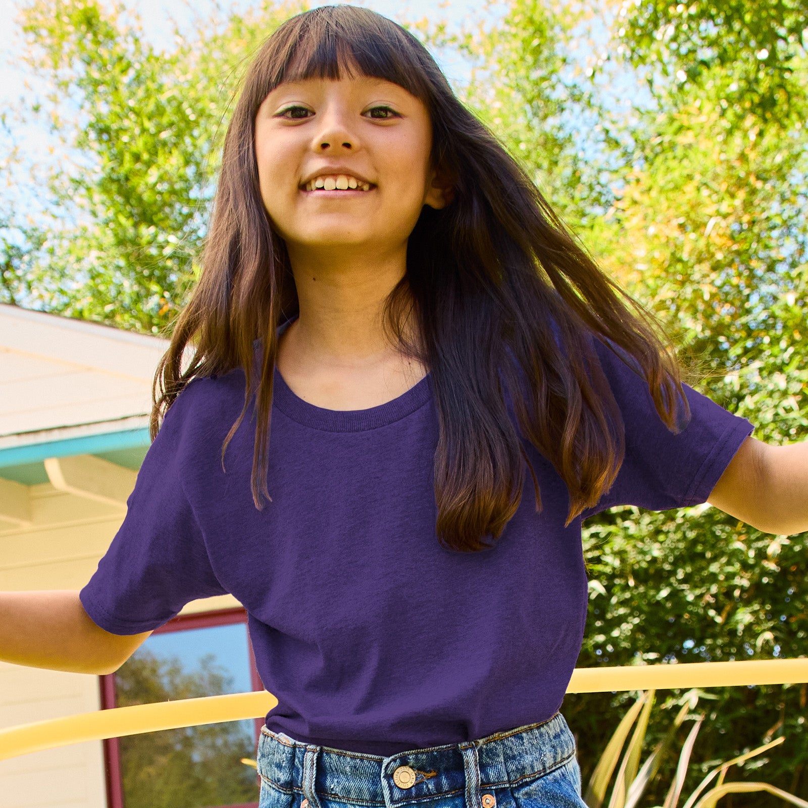 A young girl smiling while wearing a Deep Purple Jerzees Classics Youth T-Shirt outdoors.