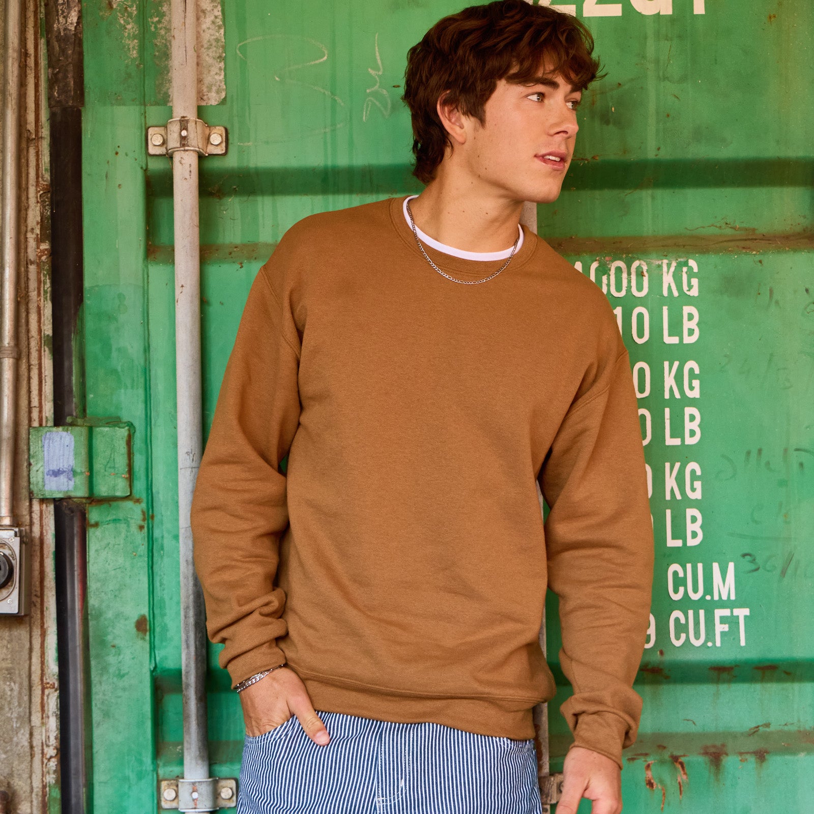 Young man modeling a Golden Pecan Jerzees NuBlend® Adult Sweatshirt against a green shipping container background.