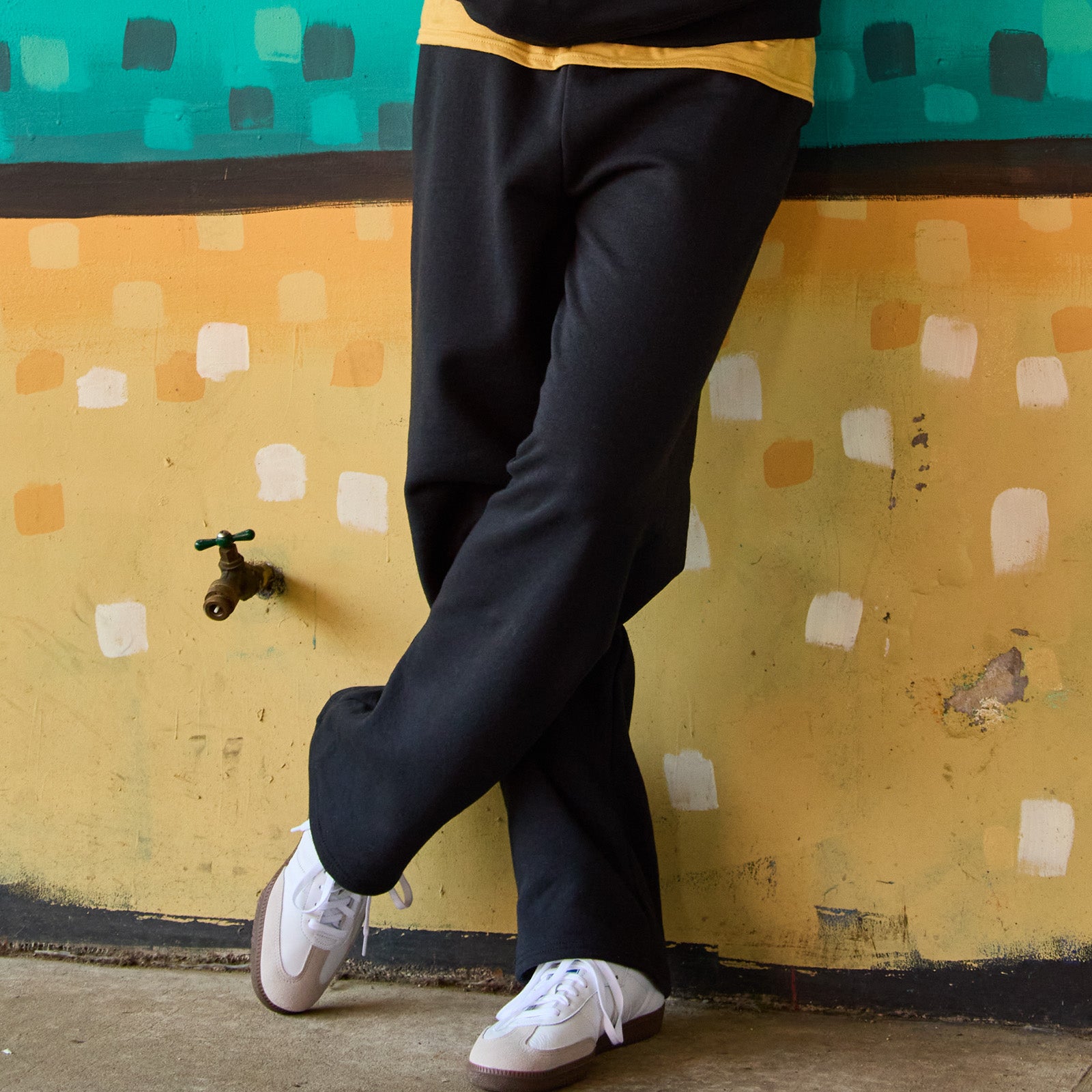 Jerzees NuBlend® black pocketed open-bottom sweatpants worn by a person leaning against a painted wall.
