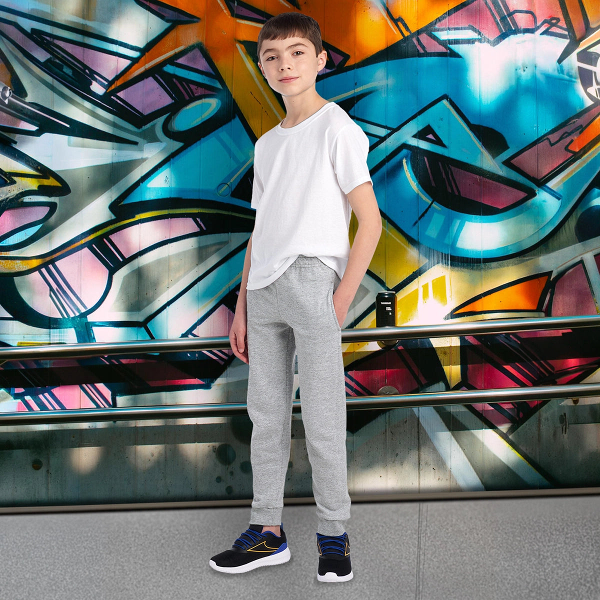Youth model wearing Jerzees NuBlend® Athletic Heather pocketed jogger sweatpants with a white t-shirt against graffiti.