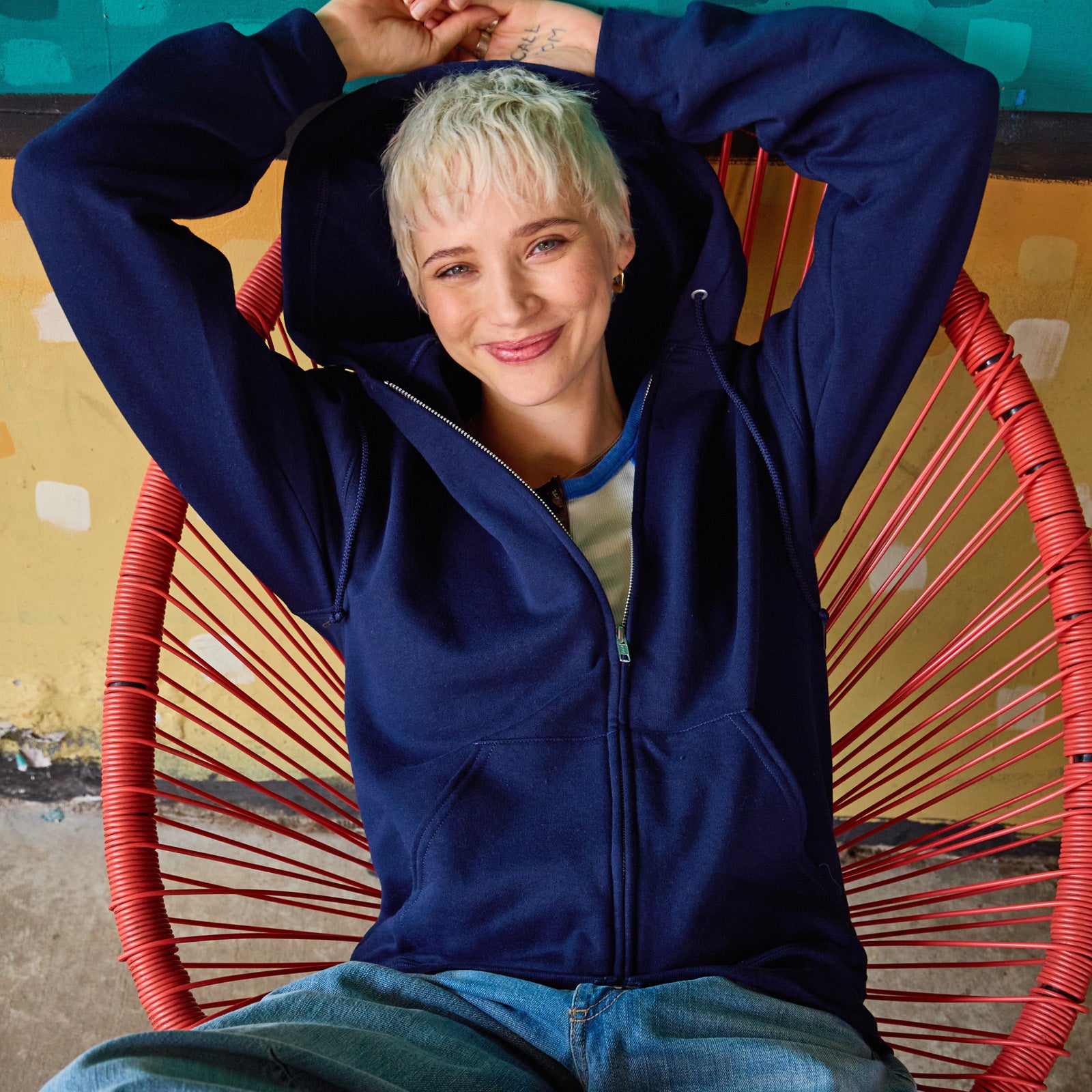 Person with blonde short hair wearing a navy Jerzees NuBlend Unisex Full-Zip Hooded Sweatshirt in a red woven chair.