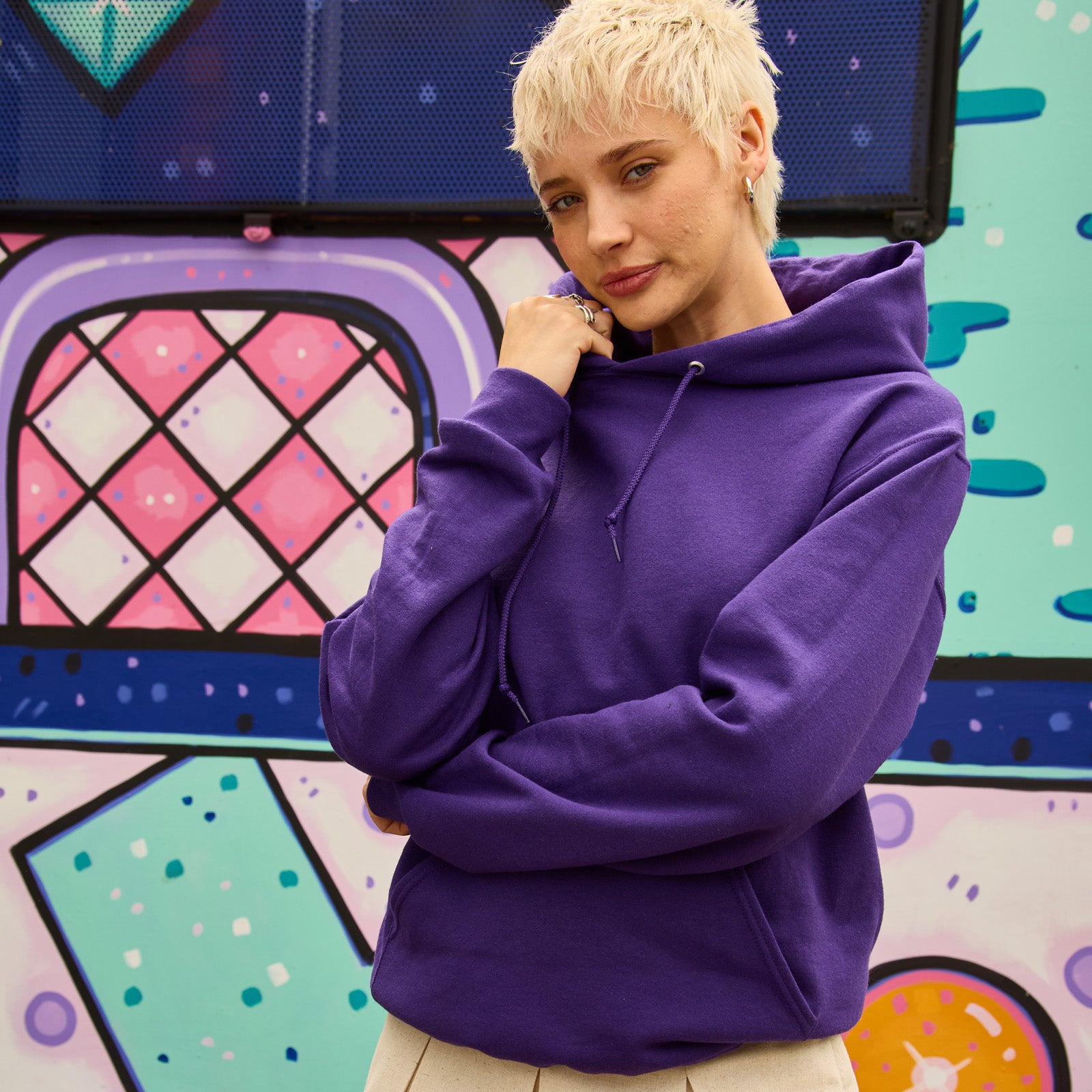 Person wearing a Jerzees NuBlend unisex hooded sweatshirt in deep purple against a colorful abstract mural background.