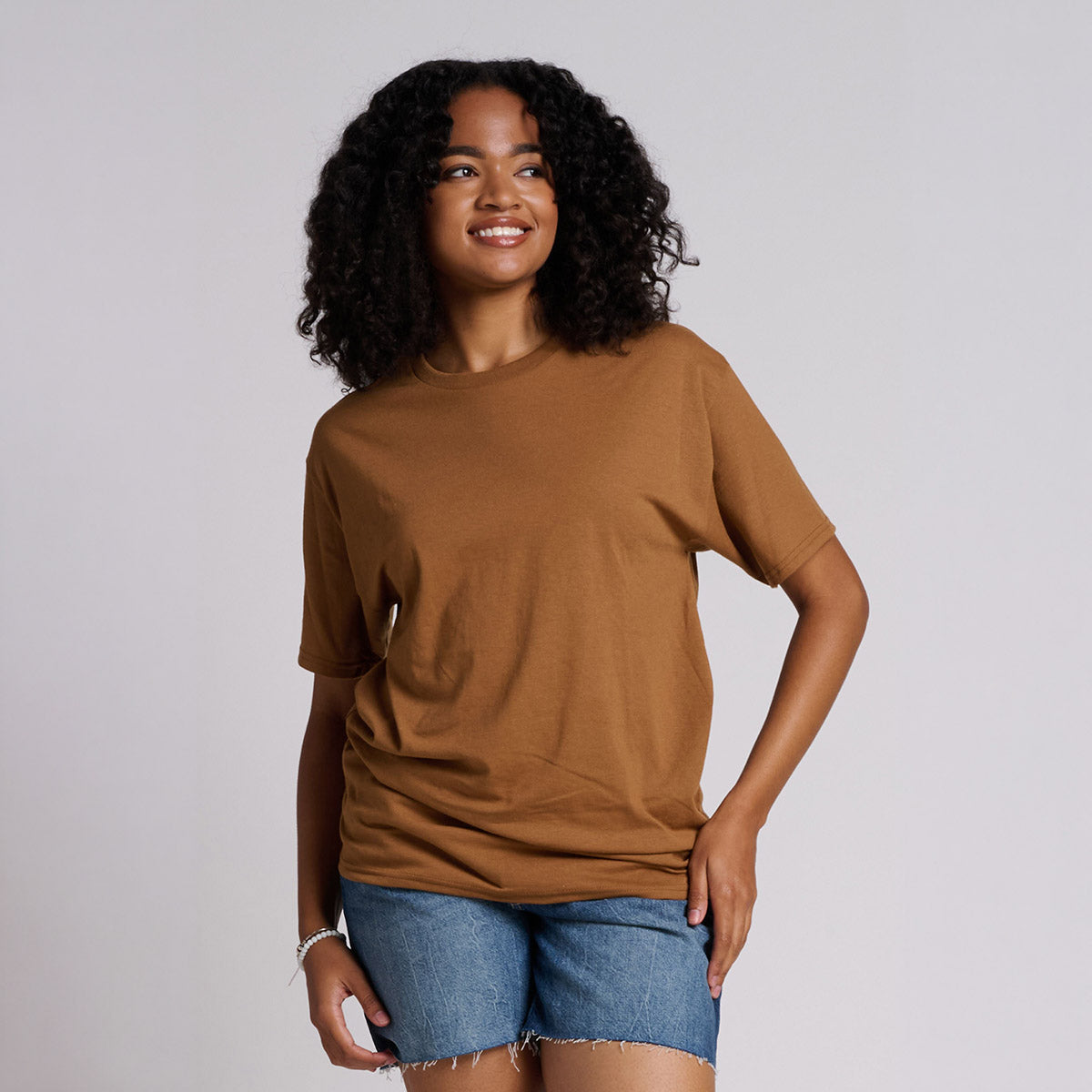 A woman wearing a Golden Pecan Jerzees Dri-Power Unisex T-Shirt with denim shorts against a light grey background.