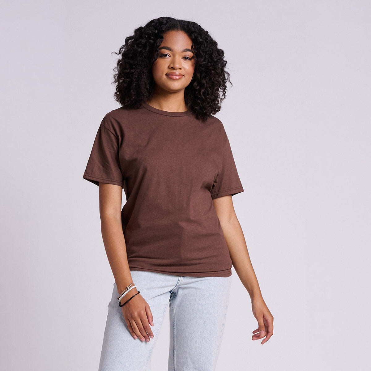 A model wearing a Jerzees Classics unisex chocolate brown t-shirt with light-wash jeans against a grey background.