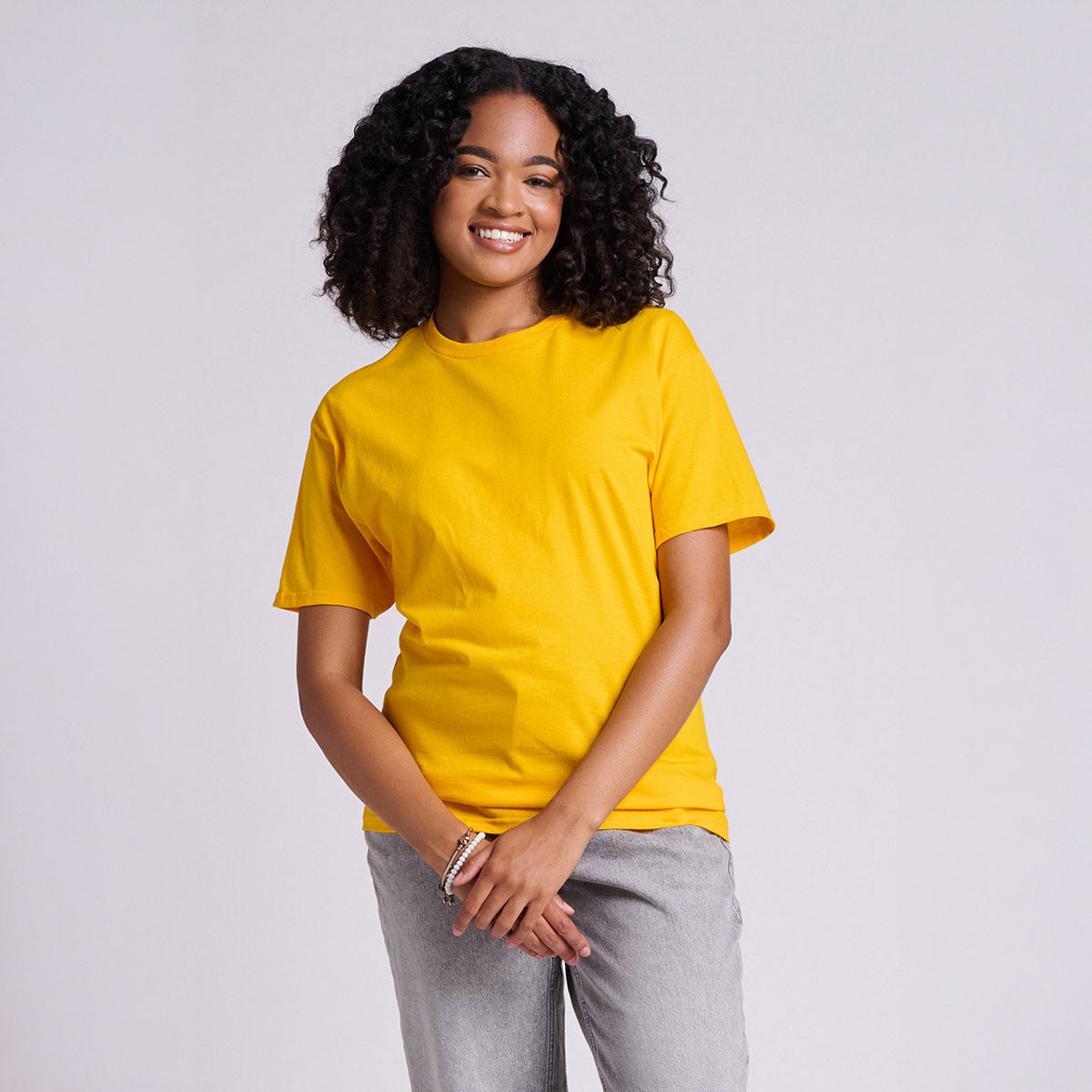 A smiling woman wearing a gold Jerzees Classics Unisex T-Shirt against a plain grey background.