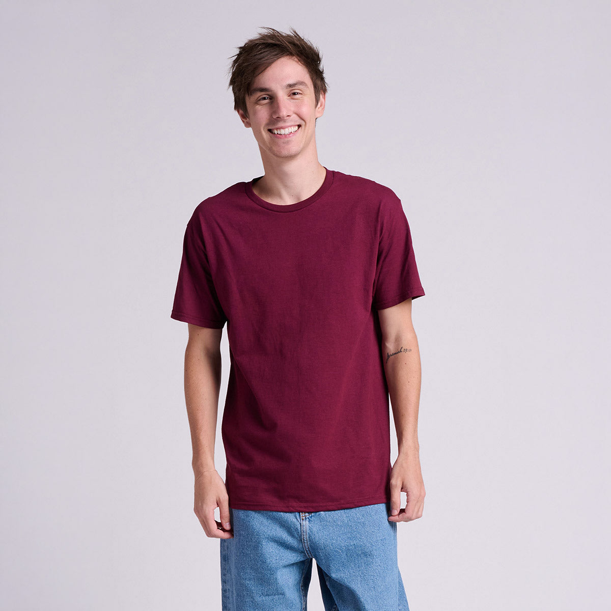 A man wearing a Jerzees Classics maroon unisex t-shirt and blue jeans, standing against a light gray background.