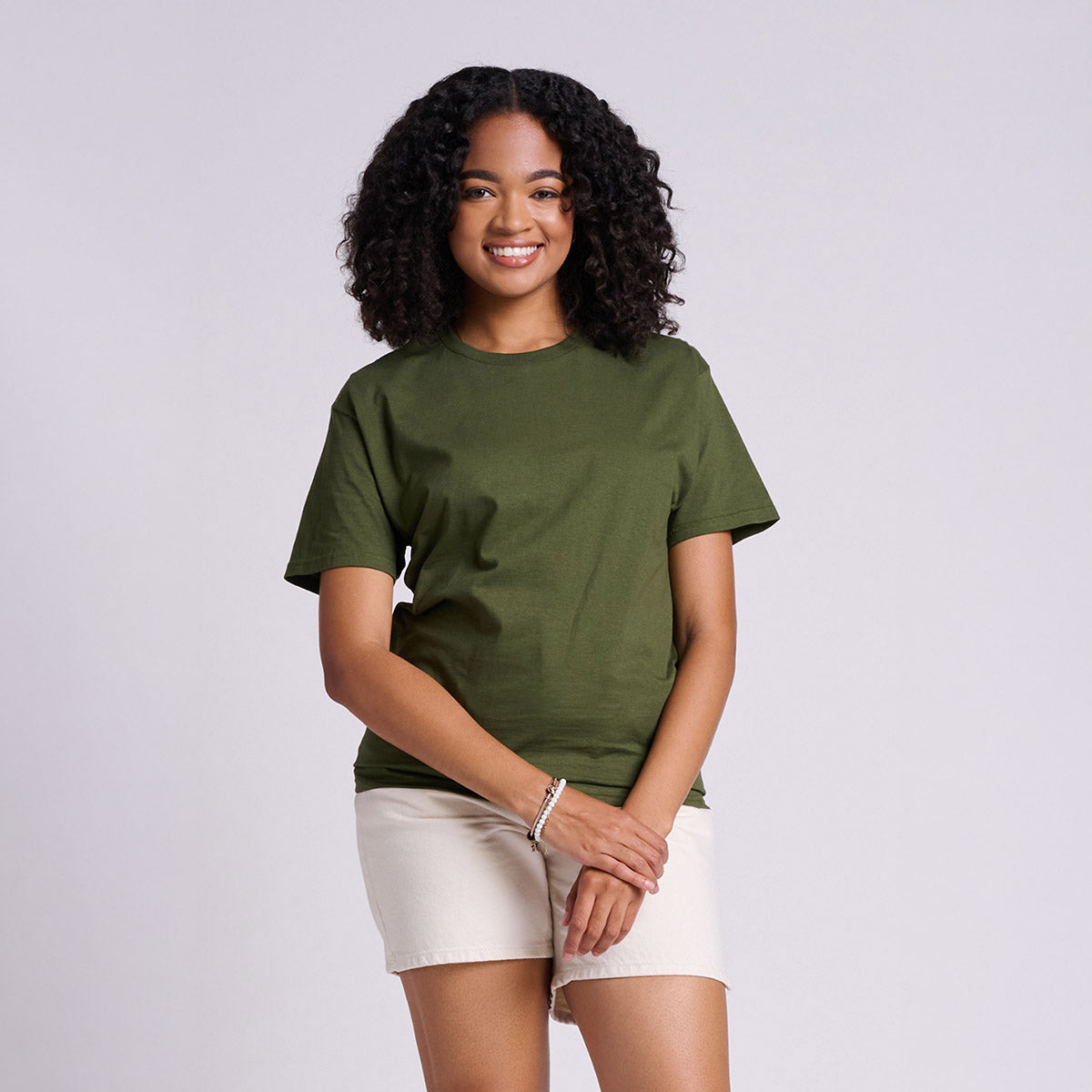 A smiling woman wearing a Military Green Jerzees Classics Unisex T-Shirt with beige shorts against a light grey background.
