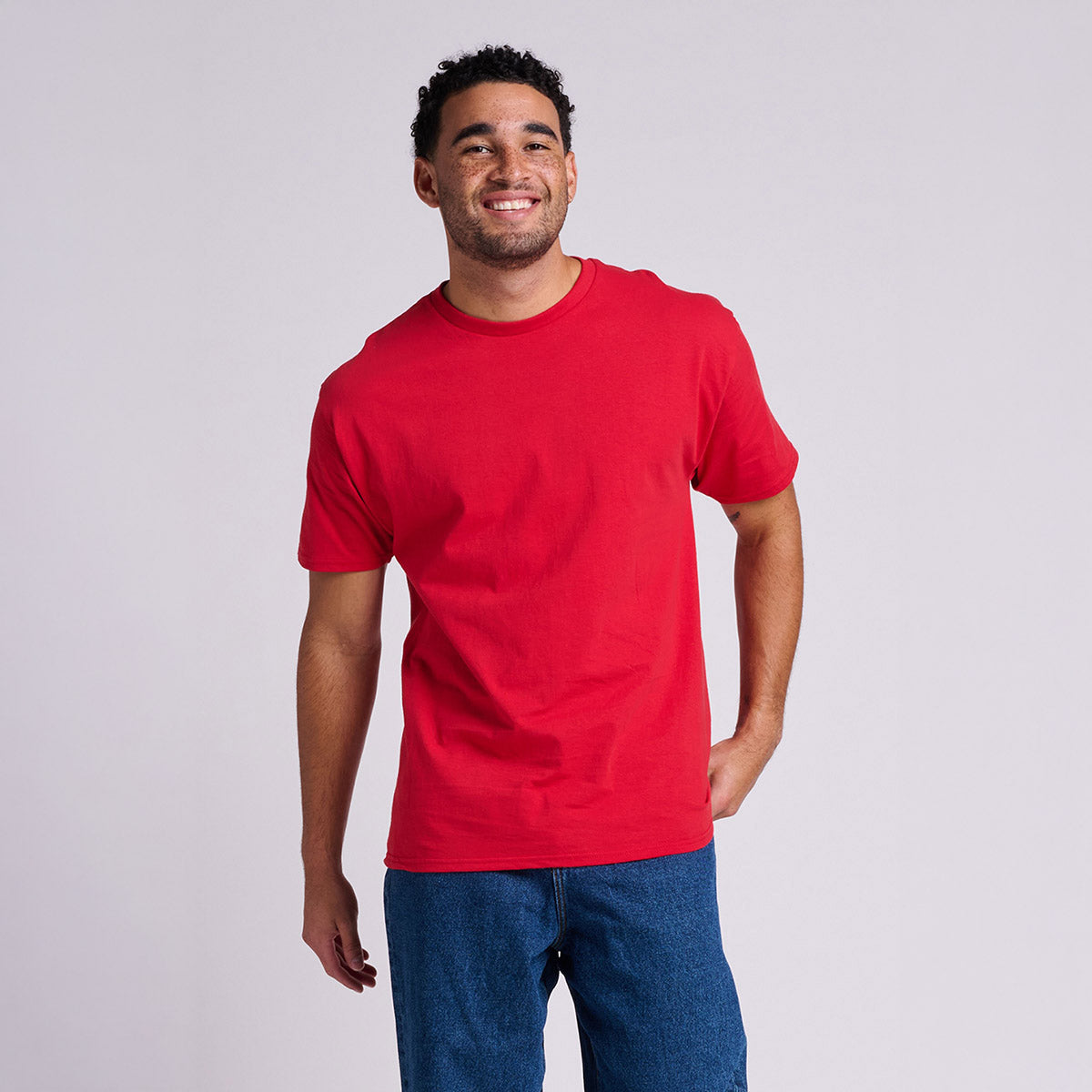 A smiling man wearing a True Red Jerzees Classics Unisex T-Shirt paired with blue jeans against a grey background.