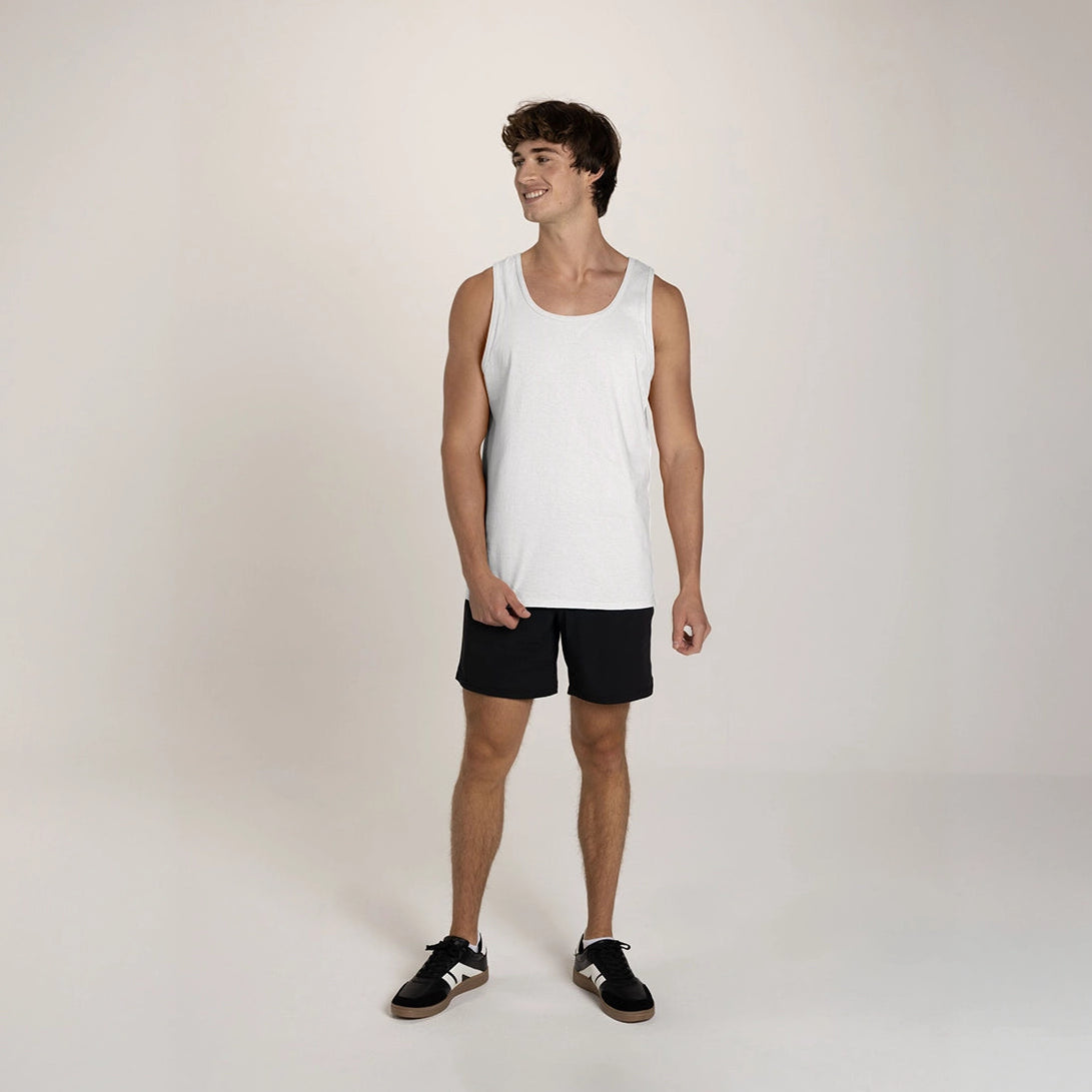 A young man wearing a white JERZEES Classics™ Unisex Tank with black shorts and sneakers against a studio background.