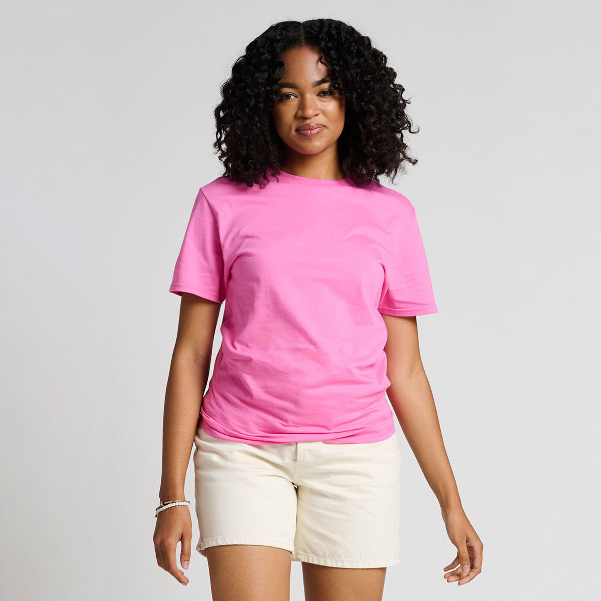 Woman with curly hair wearing a bubblegum pink Jerzees Premium Combed Ring-Spun Cotton Unisex T-Shirt with white shorts.