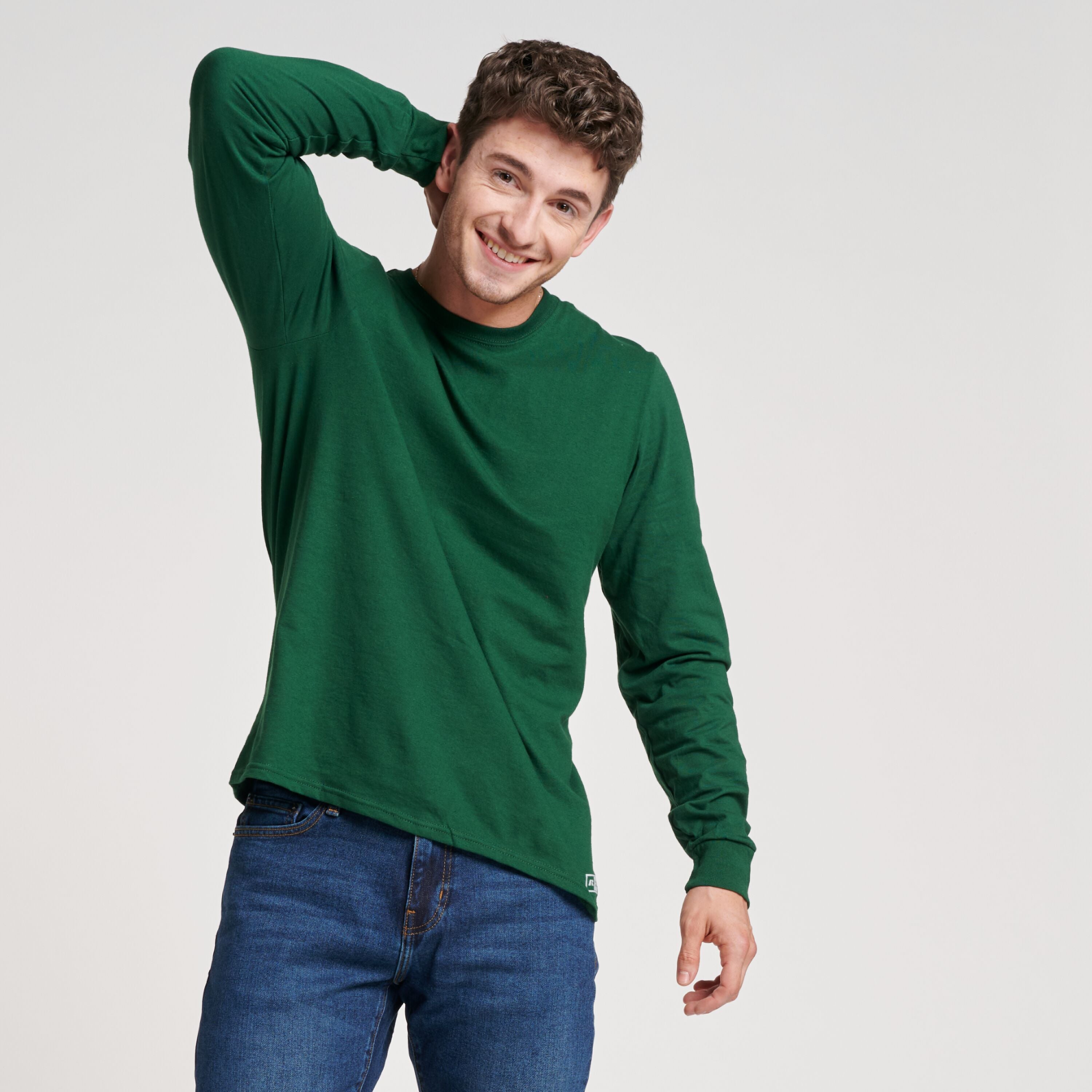 A smiling man wearing a dark green Russell Athletic DRI-POWER CVC Performance unisex long-sleeve T-shirt and blue jeans.