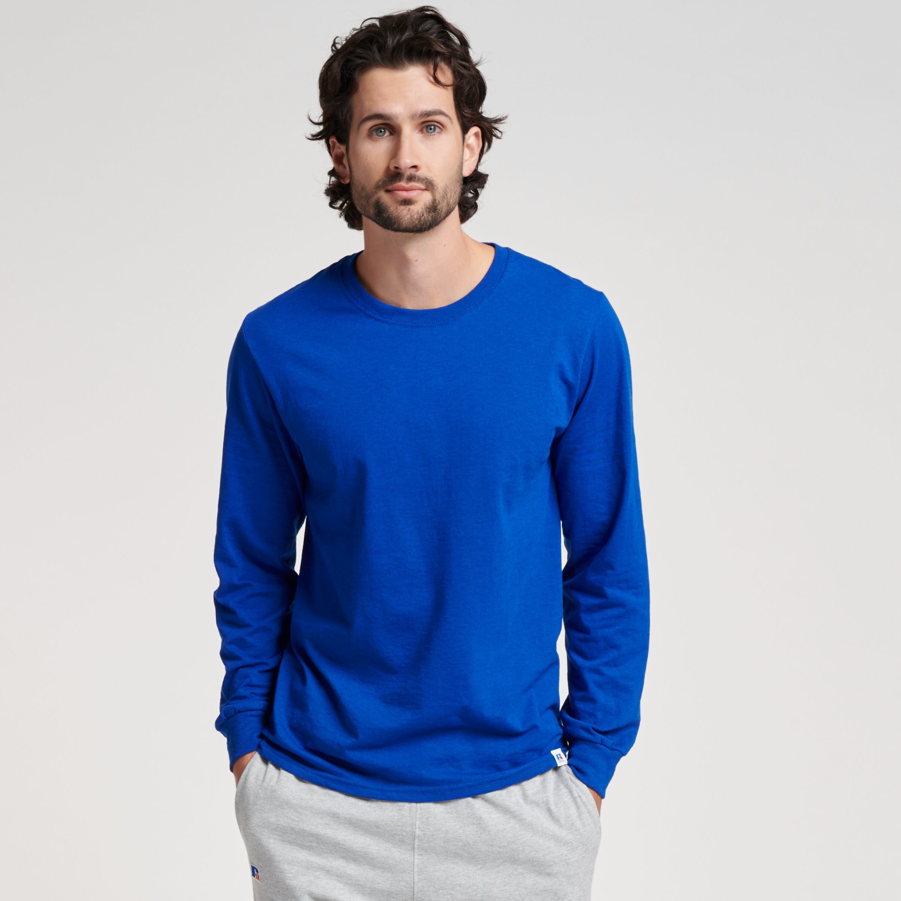 A man wearing a royal blue Russell Athletic DRI-POWER CVC Performance unisex long-sleeve T-shirt with grey sweatpants.