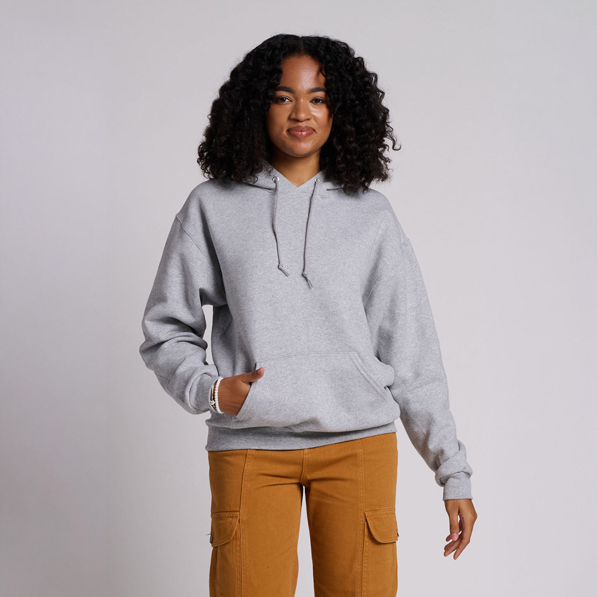 Woman with curly hair wearing an Athletic Heather Jerzees Rugged Unisex Hooded Sweatshirt with cargo pants.