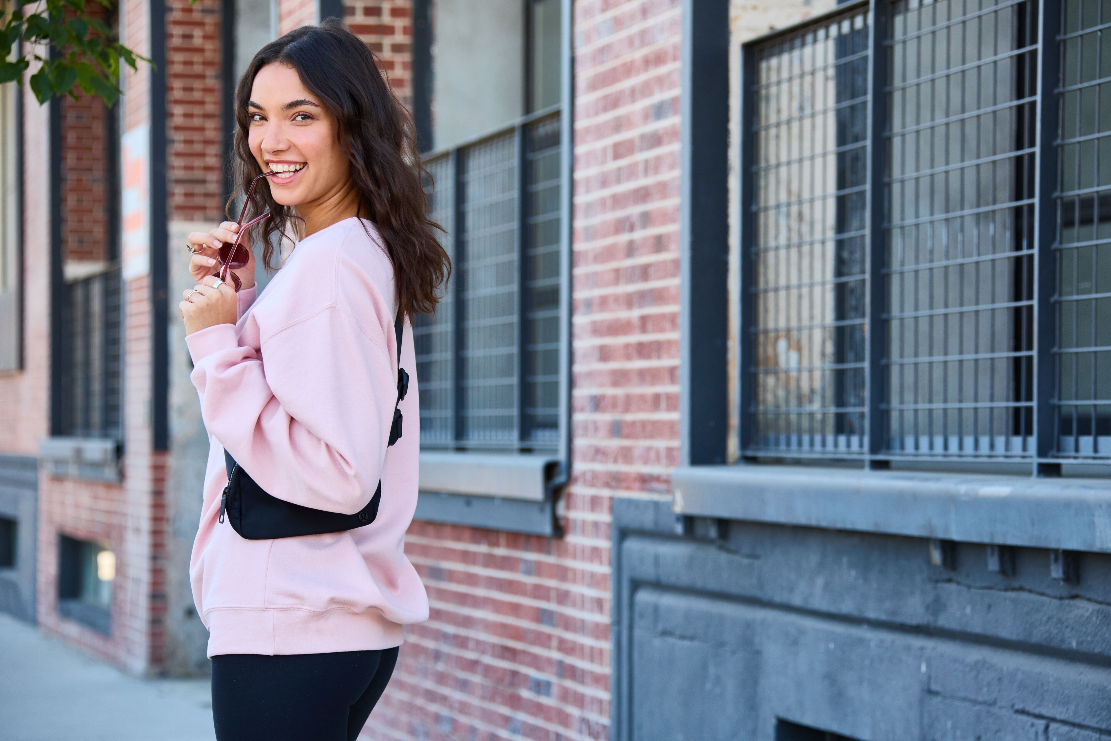 Smiling woman wearing a light pink sweatshirt and black leggings with a crossbody bag, standing in front of a brick building.