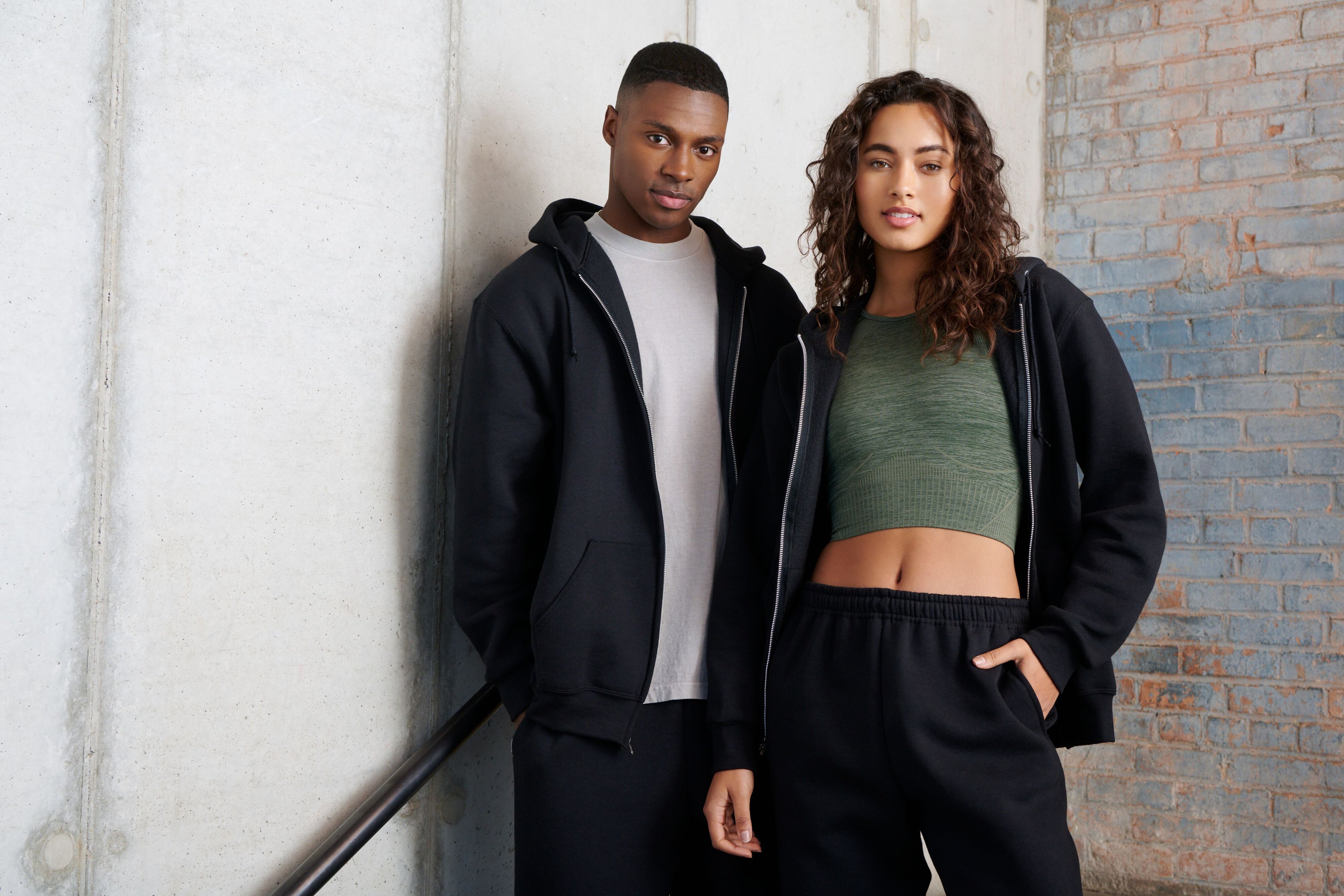 A man and woman standing together, both wearing black zip-up hoodies and matching black sweatpants.
