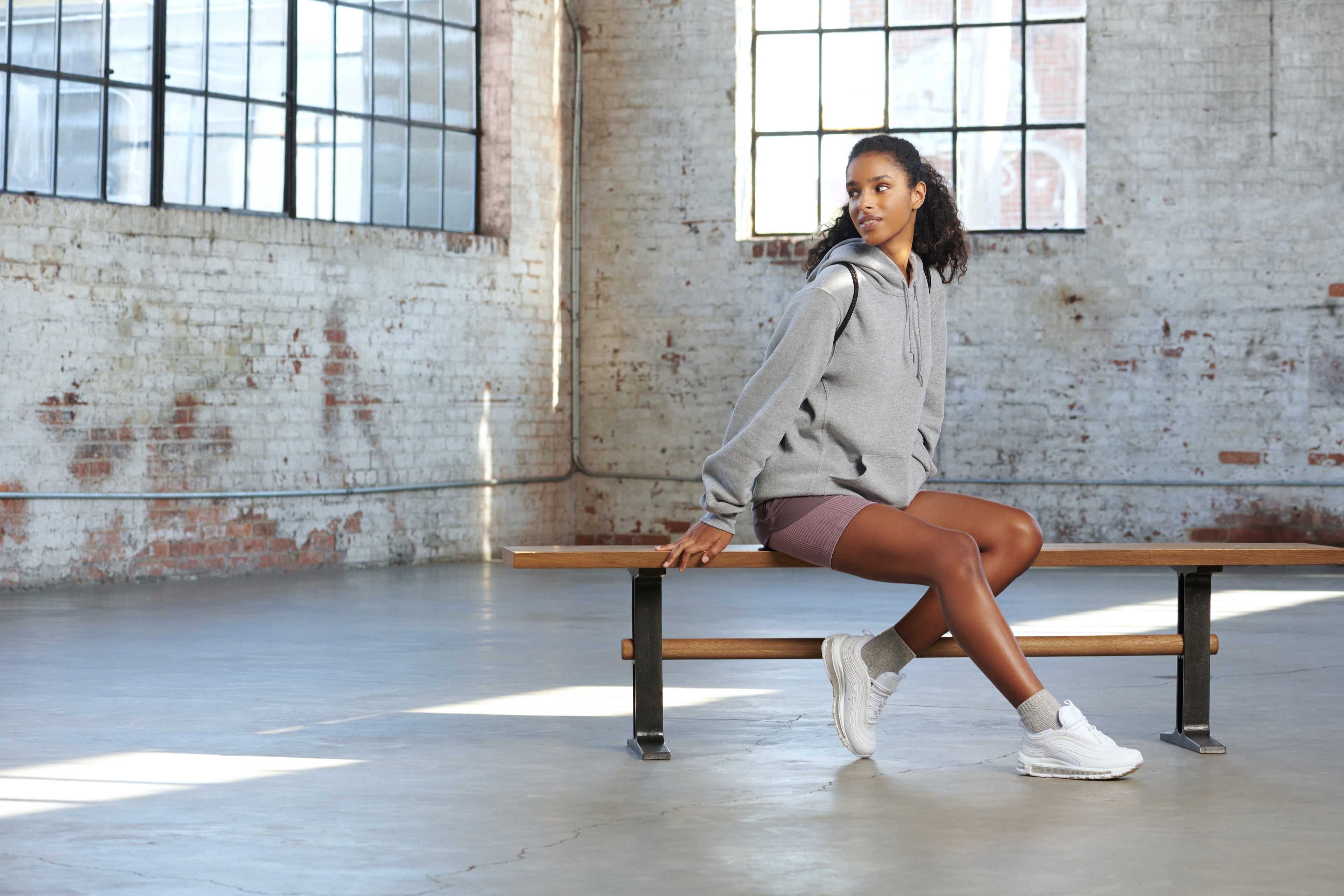 A model wearing a grey hoodie and purple shorts sits on a wooden gym bench, styled with white sneakers and grey socks.
