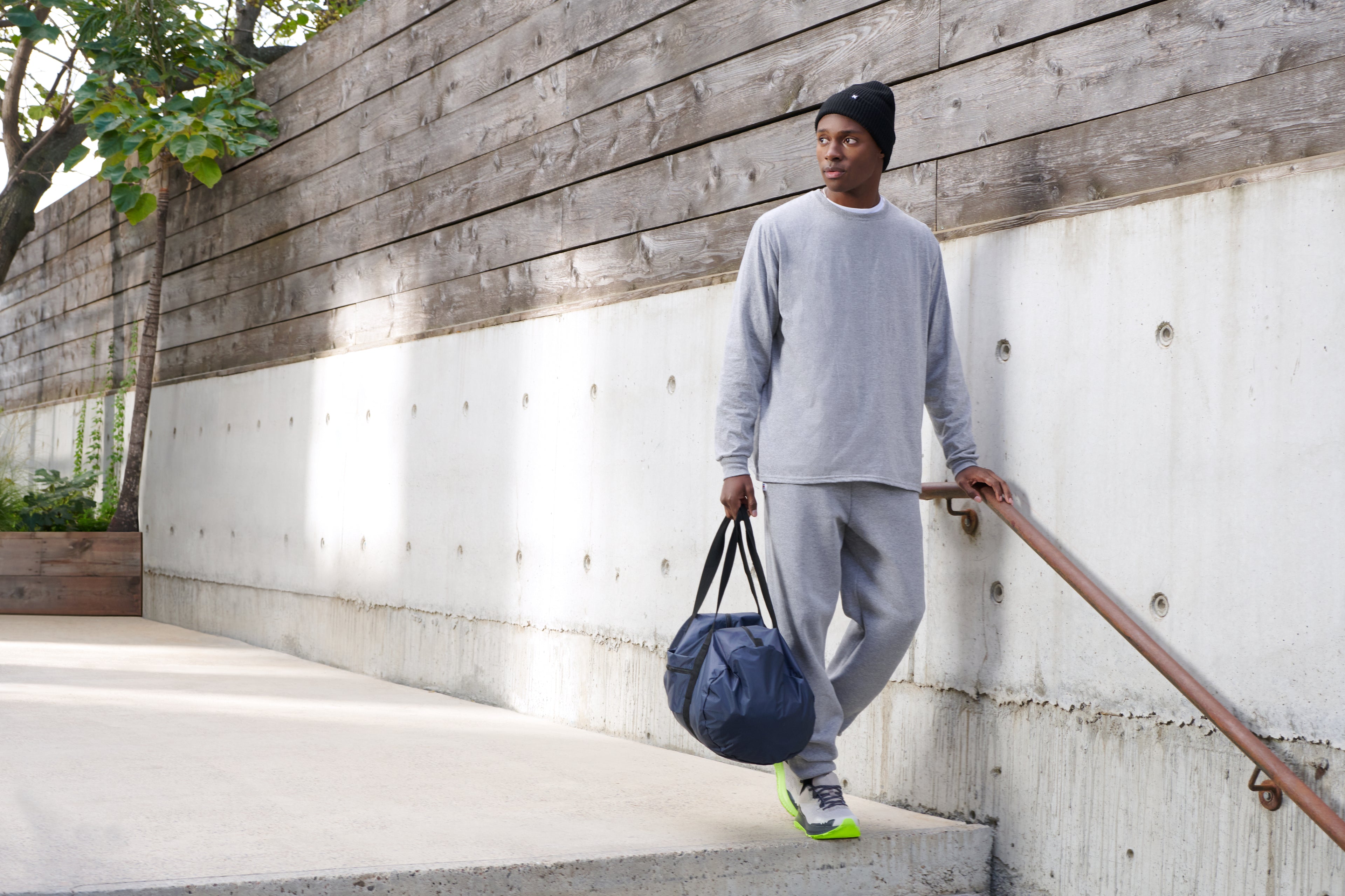 Young man in a grey sweatshirt and matching sweatpants standing on concrete stairs holding a dark blue gym duffel bag.